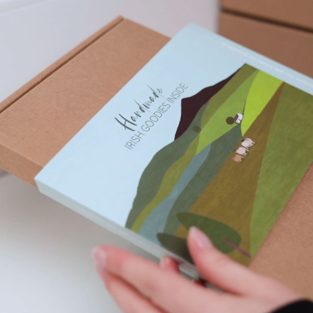 A kraft gift box wrapped in an Irish landscape sleeve featuring a blue sky, green hills and fields, an Irish cottage, sheep and trees.