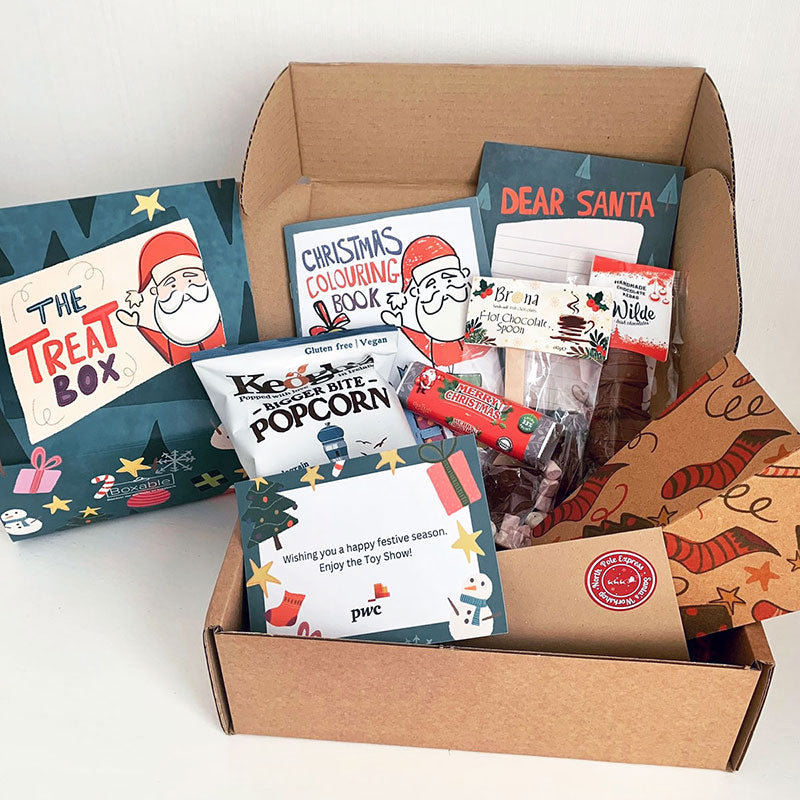 A kraft gift box filled with treats including a popcorn holder with red socks, Keogh's popcorn, a chocolate bar with a red wrapper, a Christmas colouring book, hot chocolate and a 'Dear Santa' letter.