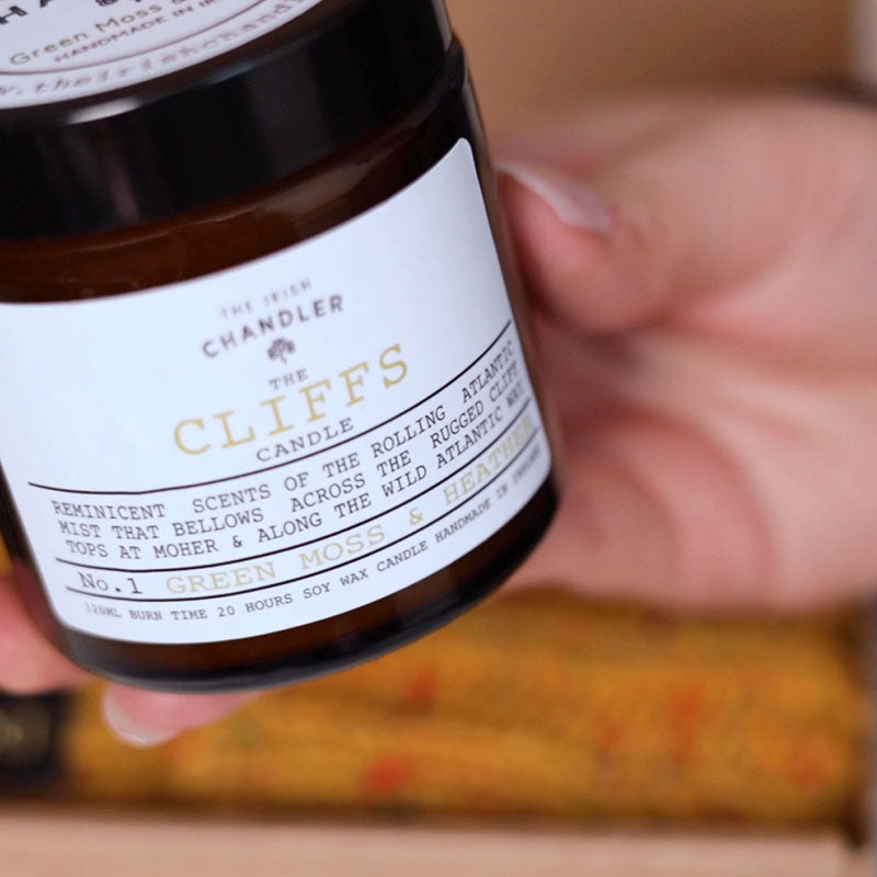 A dark brown glass candle with a white label. The text on the label reads 'The Cliffs Candle'.